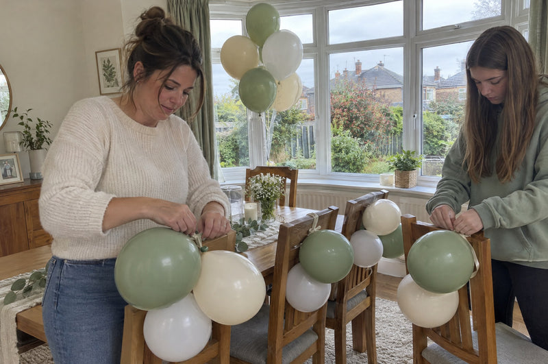 Mother and teenage daughter tying sage green and cream helium balloons to dining chairs for a baby shower